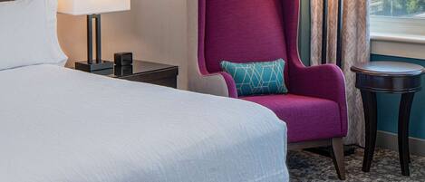 A hotel room with a large bed, a purple armchair, a side table with a lamp, a framed picture on the wall, and a view of the outdoors through a window.