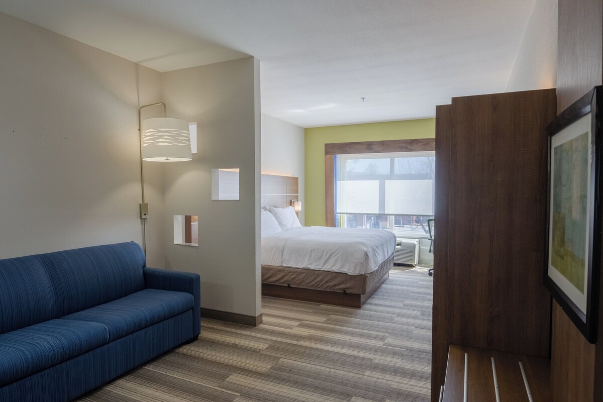 Quarto de hotel com um sofá azul, uma cama com roupa de cama branca, uma janela grande e um quadro na parede.