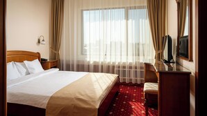 A hotel room with a bed, a desk with a television, a chair, a window with curtains, and a wall-mounted lamp. - Traian Braila (Braila)