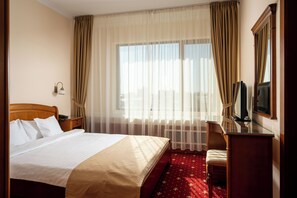 A hotel room with a bed, a desk with a television, a chair, a window with curtains, and a wall-mounted lamp. - Traian Braila (Braila)