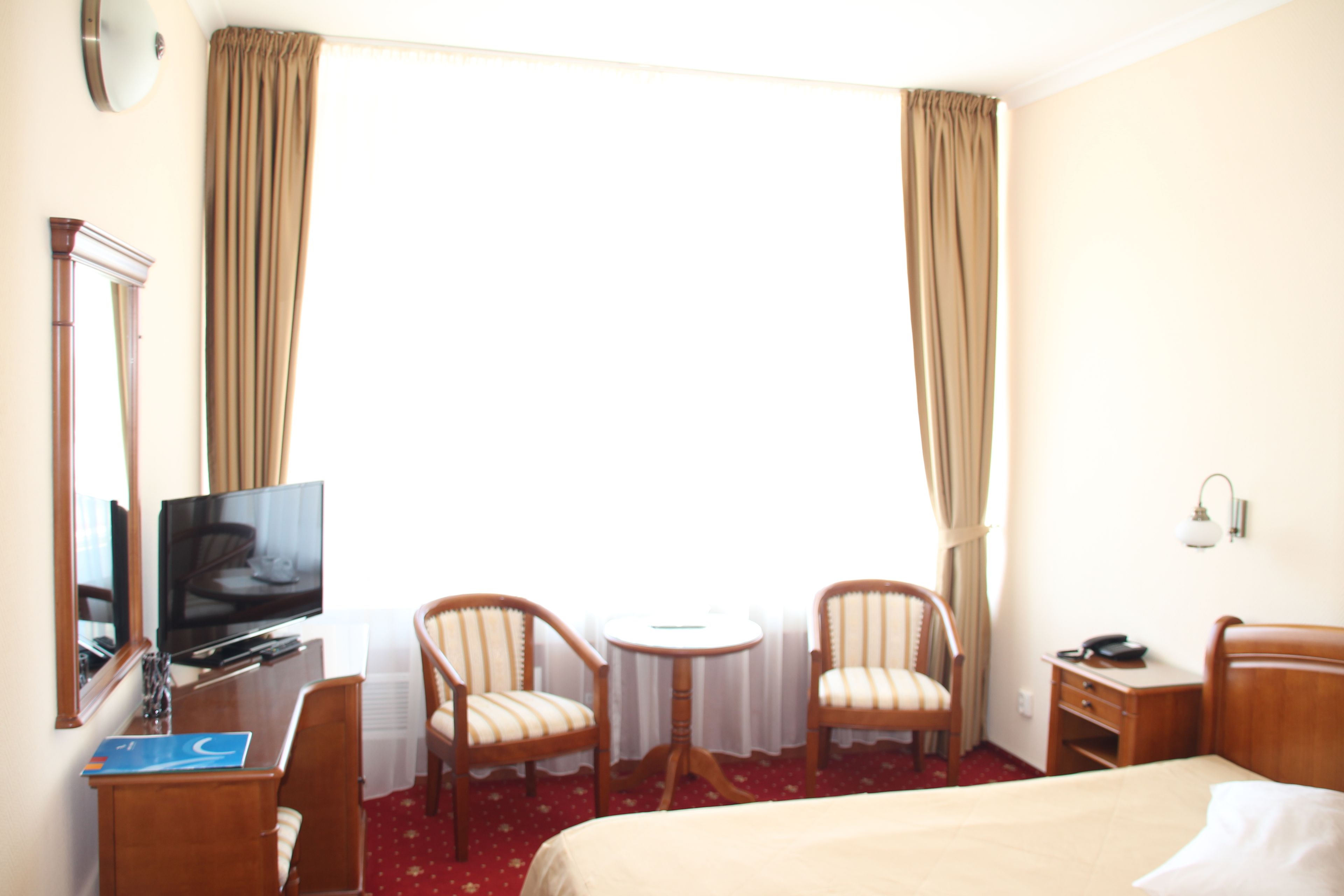 Habitación de hotel con cama, escritorio con televisor, dos sillas, una mesita y ventana con cortinas.