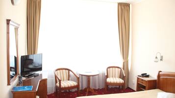 Habitación de hotel con cama, escritorio con televisor, dos sillas, una mesita y ventana con cortinas.