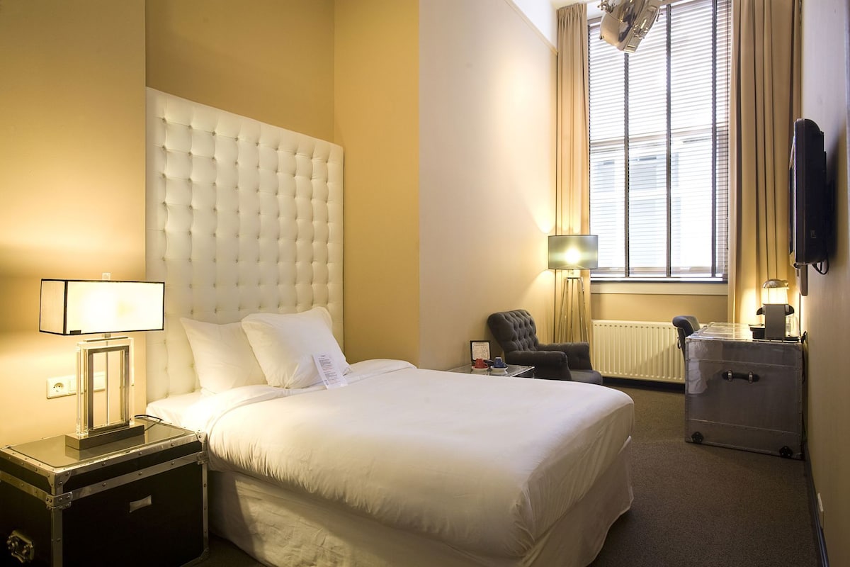 Habitación de hotel con una cama grande, una mesita de noche con lámpara, una silla, un escritorio con computadora y una ventana con cortinas.