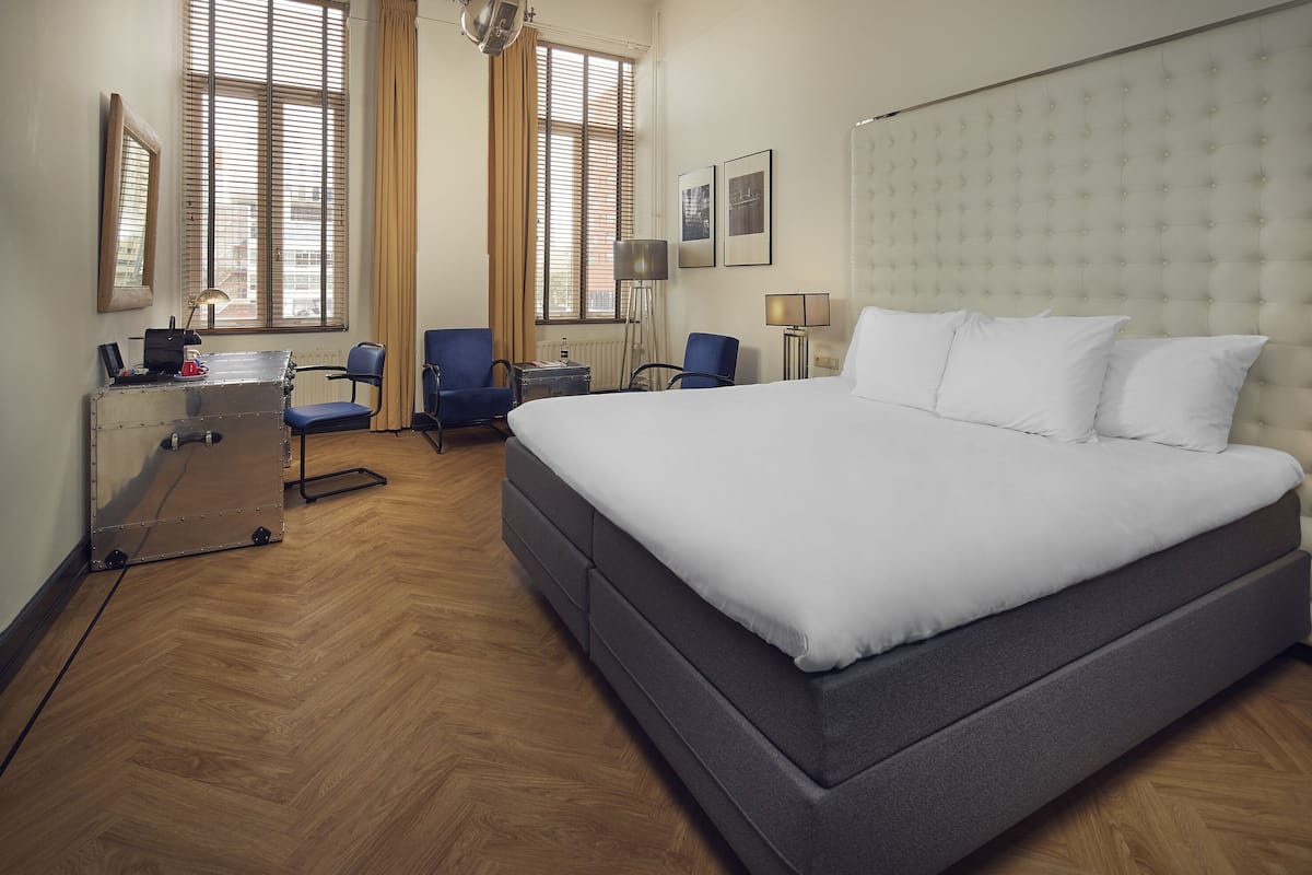 A hotel room with a large bed, two chairs, a desk, and a window with blinds.