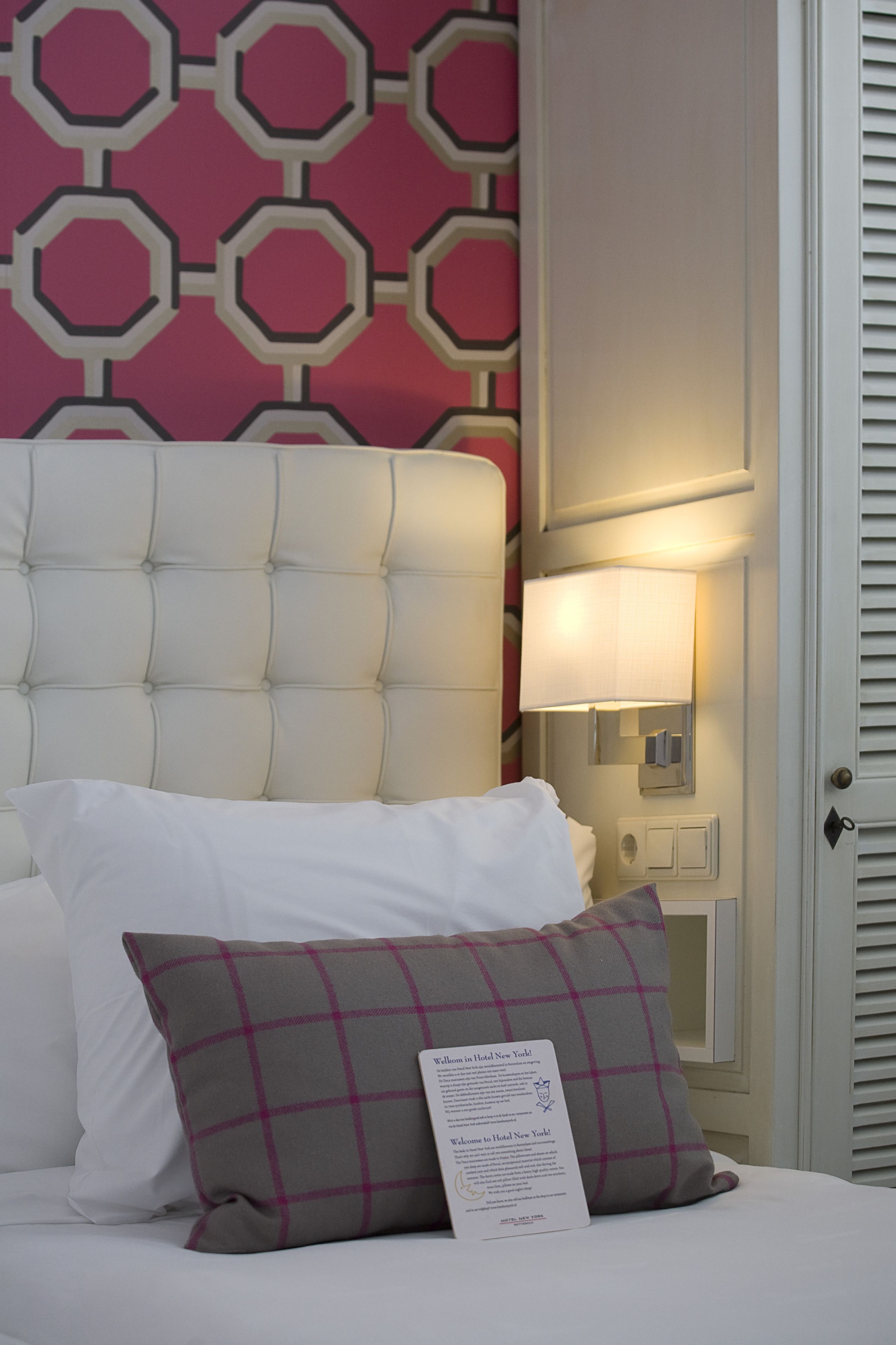 Ein Hotelzimmer mit einer gebügelten Kopfstütze, einer Nachttischlampe, einem Kissen und einem Willkommensschild.