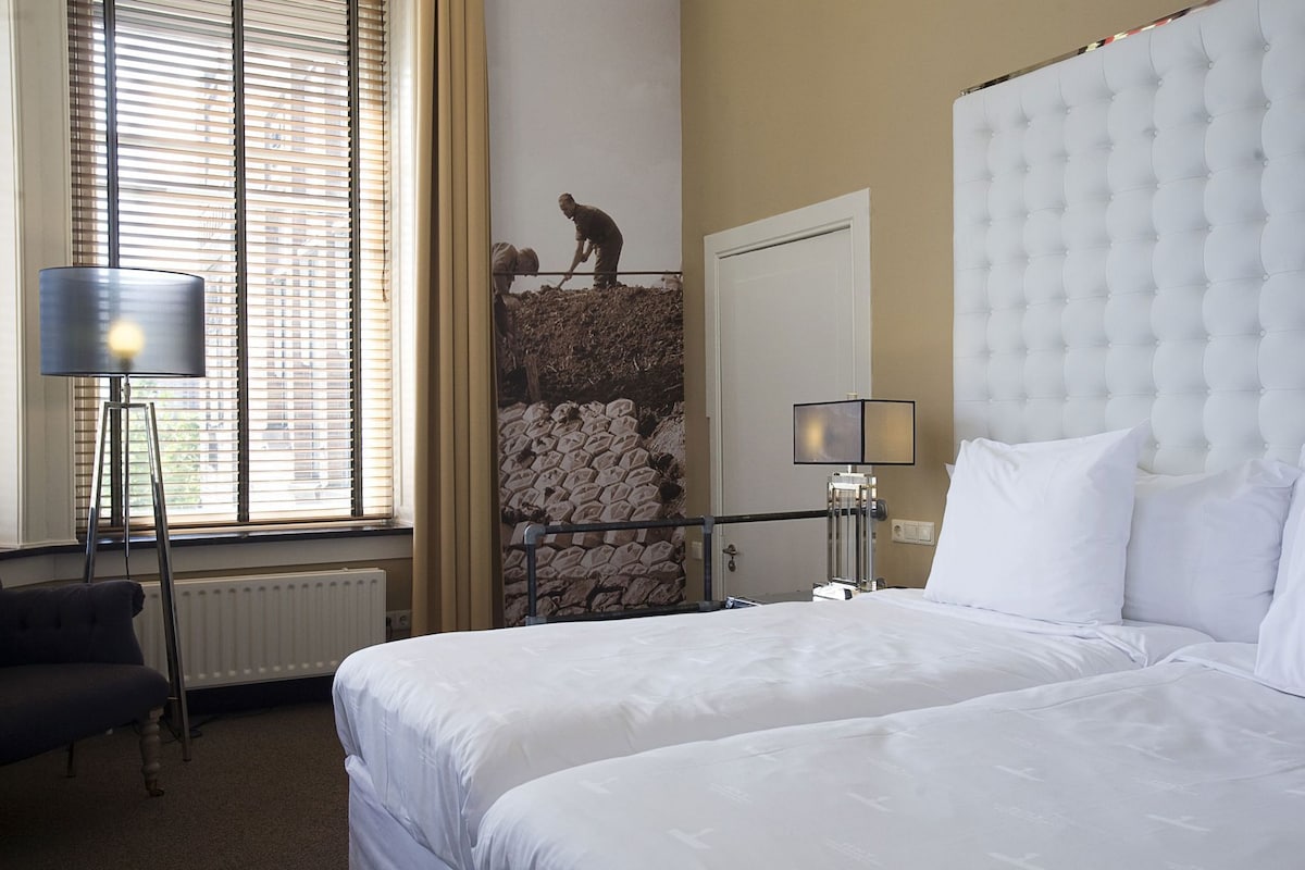 Habitación de hotel con cama, lámpara, radiador, ventana con persianas y pared de piedra con estatua.
