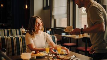 Un coin repas avec une table dressée pour deux personnes, un canapé et une fenêtre avec des stores.
