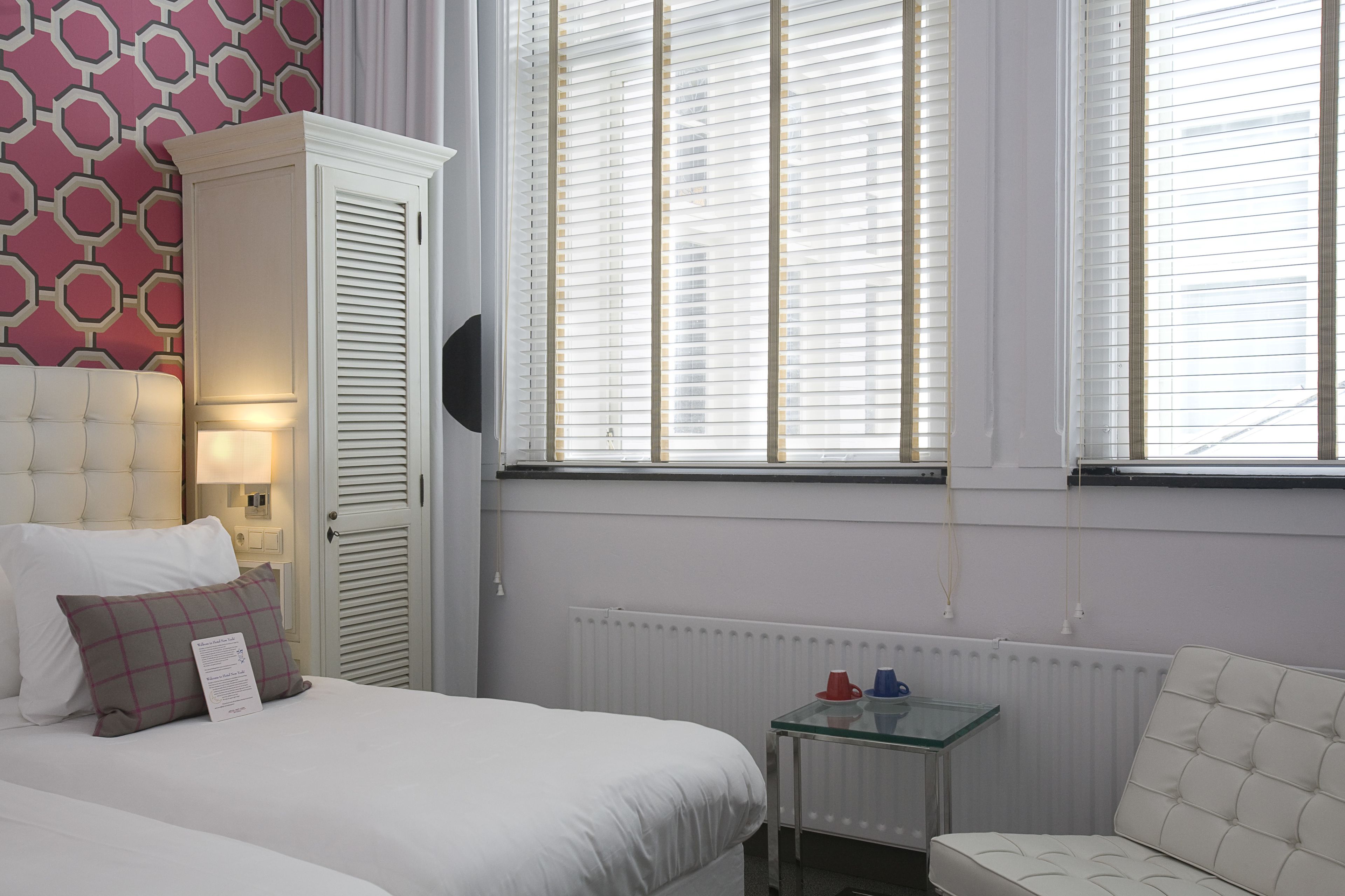 A hotel room with a bed, a white wardrobe, a glass table with two cups, and a window with blinds.