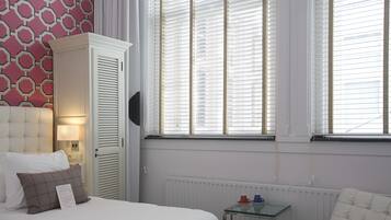 A hotel room with a bed, a white wardrobe, a glass table with two cups, and a window with blinds.