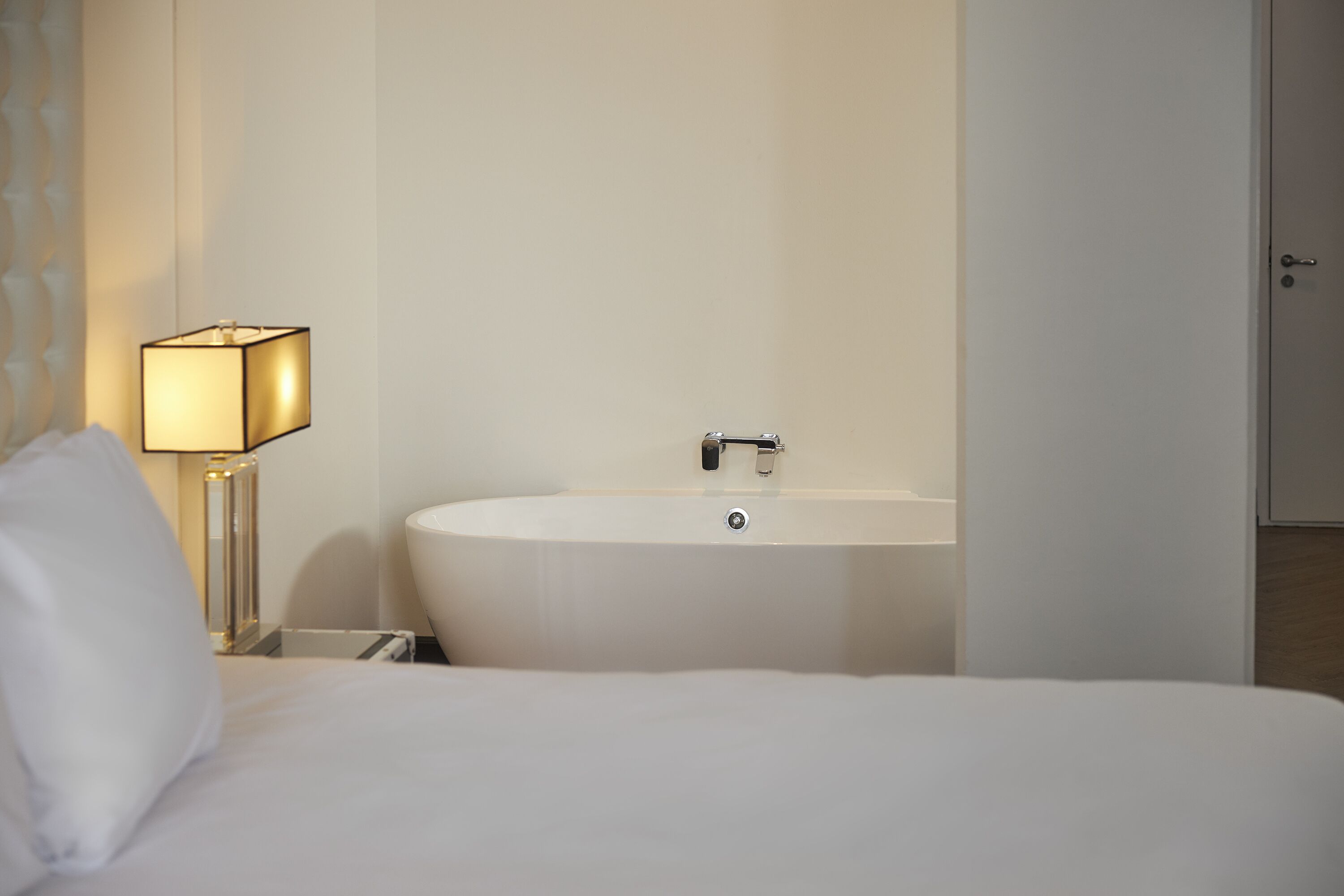 Una habitación de hotel con cama, lámpara de noche y una bañera blanca.