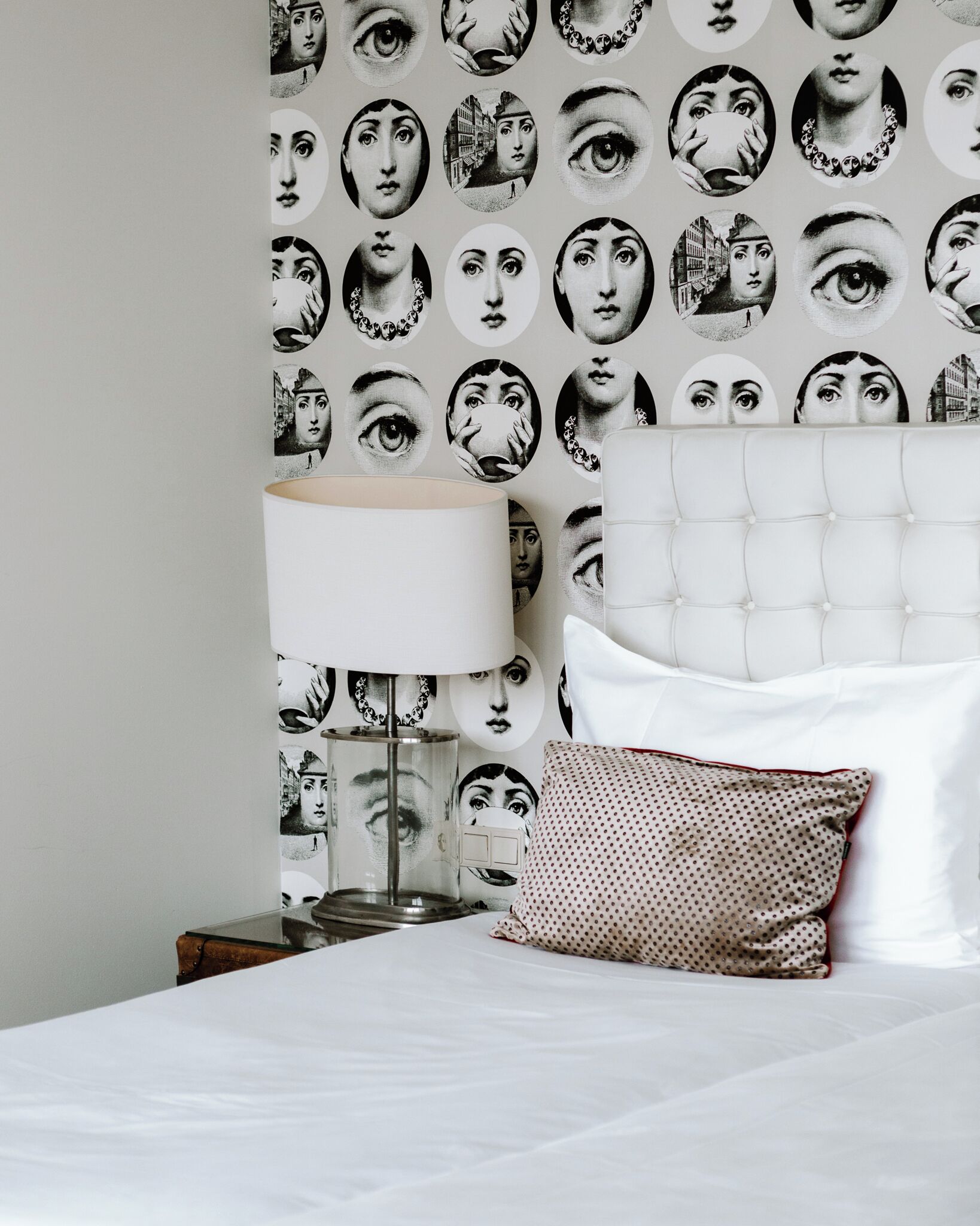 A neatly made bed with white linens, a white tufted headboard, a patterned pillow, a bedside lamp, and a wall with a repeating pattern of faces.