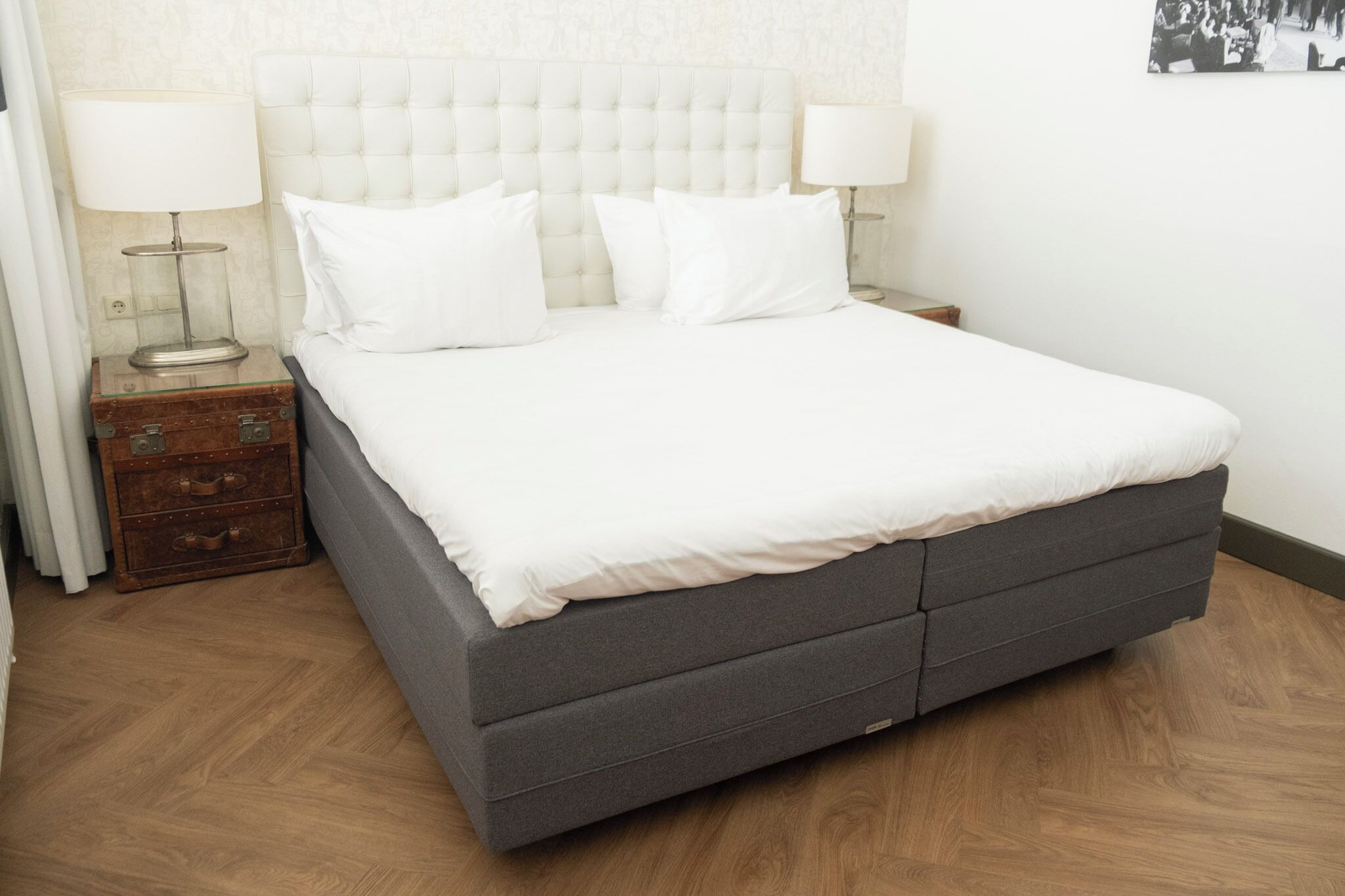 Una cama bien hecha con base gris y ropa de cama blanca, flanqueada por dos mesitas de noche con lámparas, en una habitación con piso de madera y un papel tapiz estampado.
