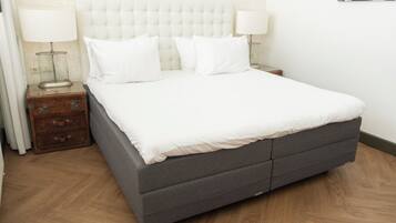 Una cama bien hecha con base gris y ropa de cama blanca, flanqueada por dos mesitas de noche con lámparas, en una habitación con piso de madera y un papel tapiz estampado.