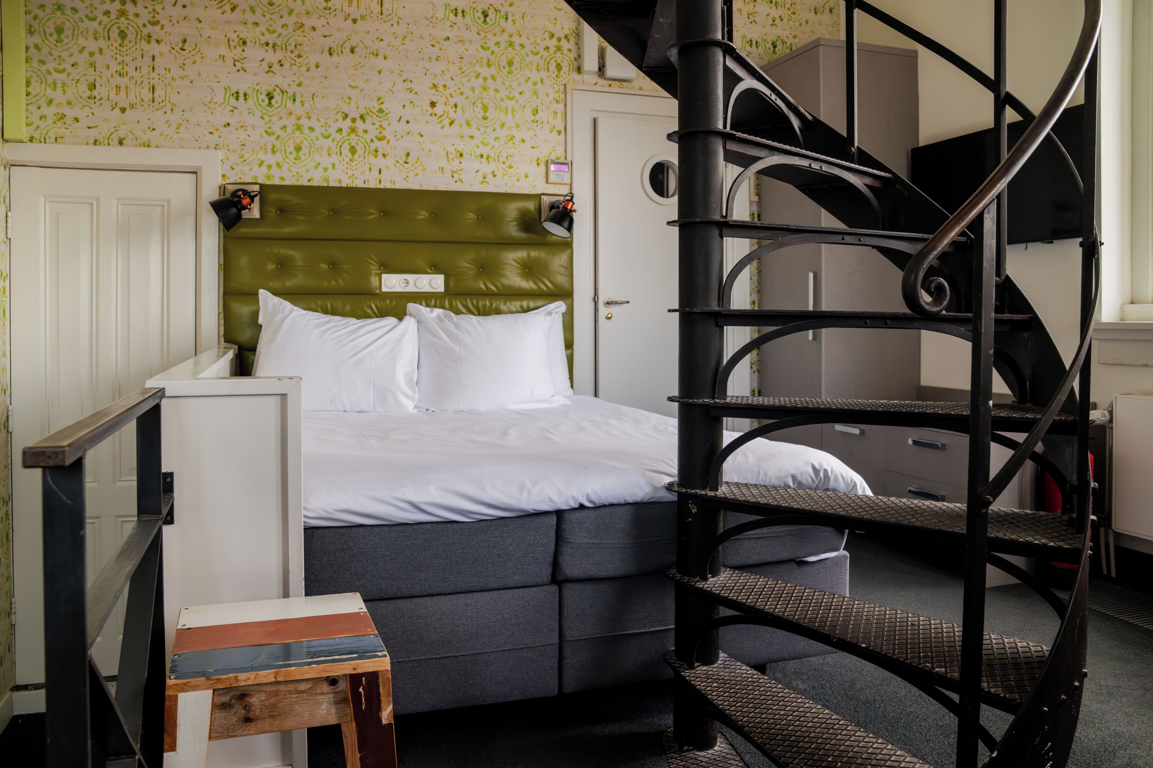 Una habitación con una cama, una pequeña mesa de madera, un cabecero tapizado en verde, una escalera espiral negra y un papel tapiz estampado.