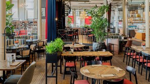 Un restaurante con varias mesas y sillas, algunas con cojines rojos. Hay plantas en macetas y un candelabro.