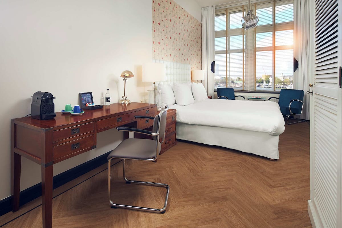 A hotel room with a wooden desk, a bed, a chair, and a window with blinds.