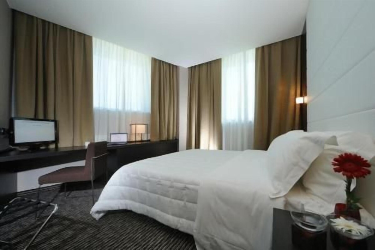 A hotel room with a bed, a desk with a computer, a chair, and a window with curtains.