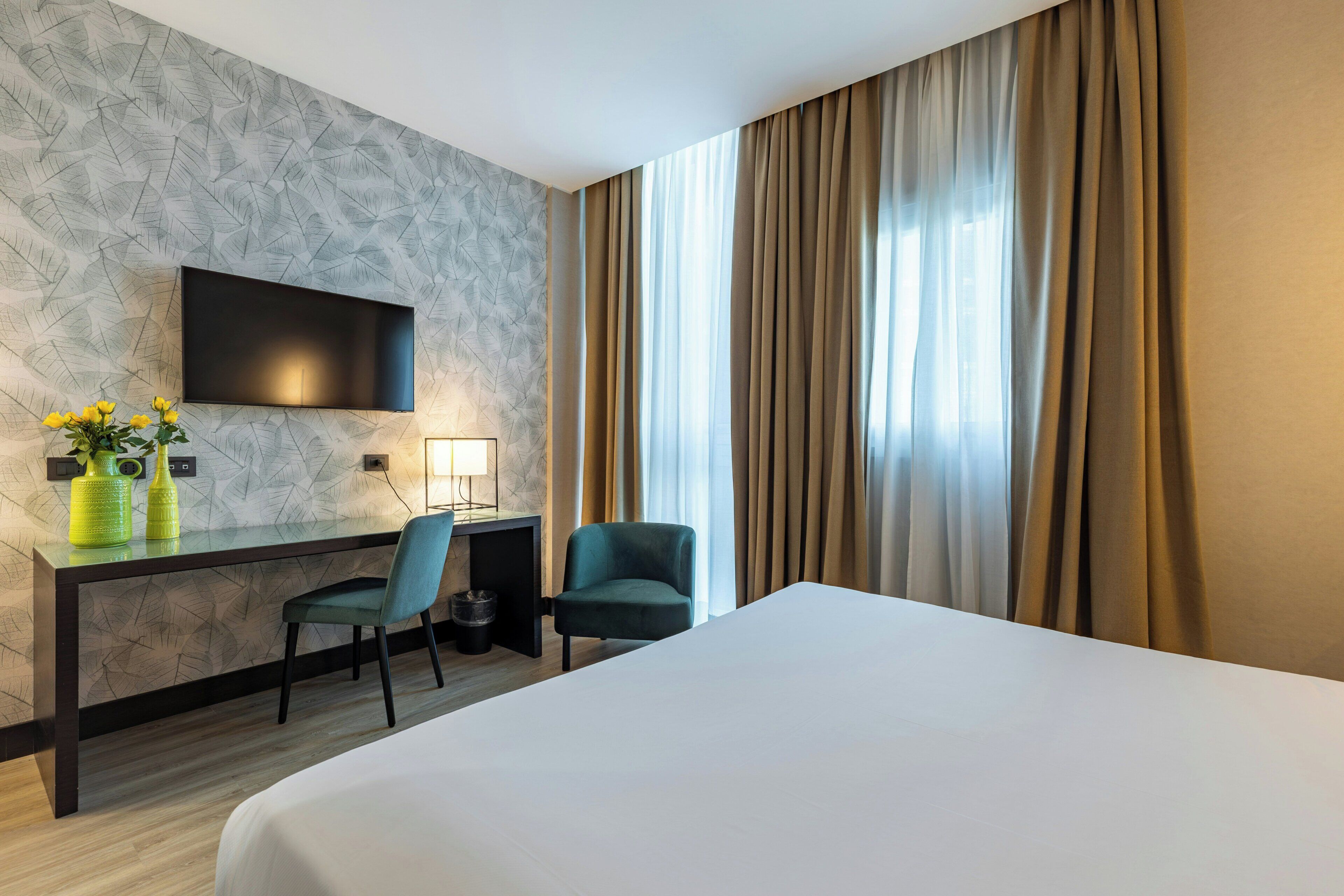 Une chambre d’hôtel comprenant un lit, un bureau avec une chaise, un téléviseur fixé au mur et une fenêtre avec des rideaux.