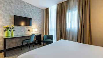 Une chambre d’hôtel comprenant un lit, un bureau avec une chaise, un téléviseur fixé au mur et une fenêtre avec des rideaux.