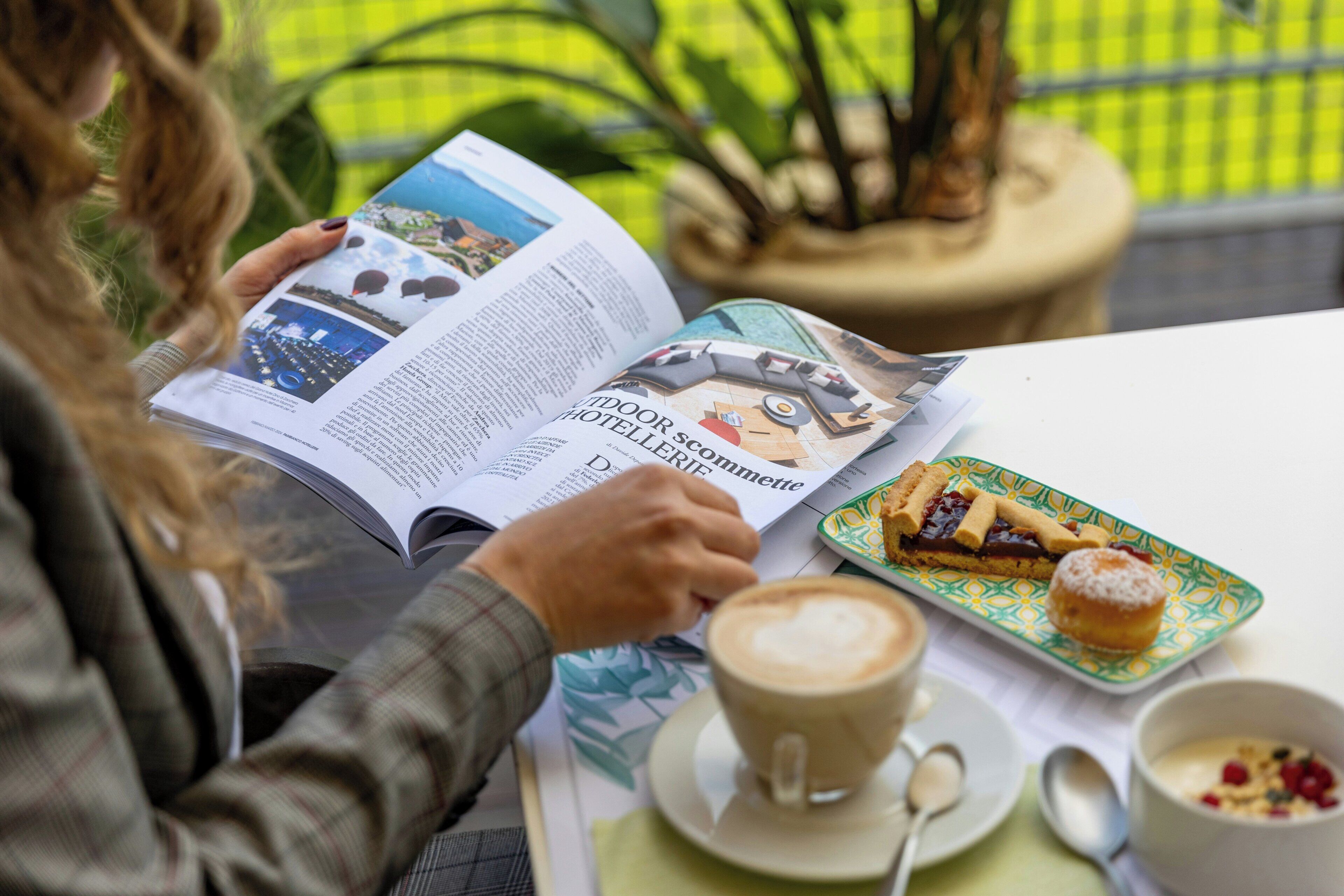 Una persona leyendo una revista en una mesa con un plato de pasteles y una taza de café.