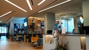 A modern hotel lobby with wooden flooring, a reception desk, seating area, and a kitchen in the background.