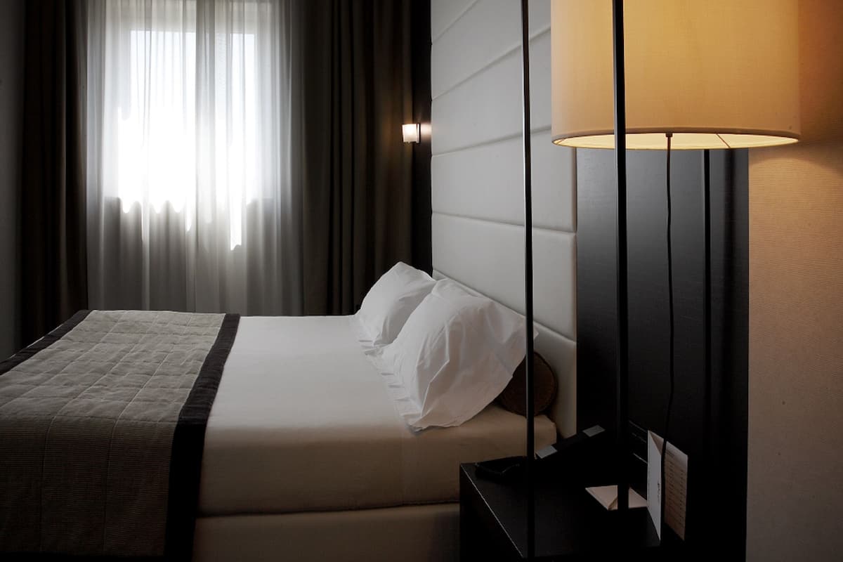 Een hotelkamer met een bed, nachtkastje, lamp en raam met gordijnen.
