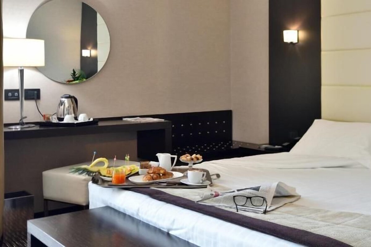 A hotel room with a bed, bedside table, and a breakfast tray with croissants, coffee, and fruit.