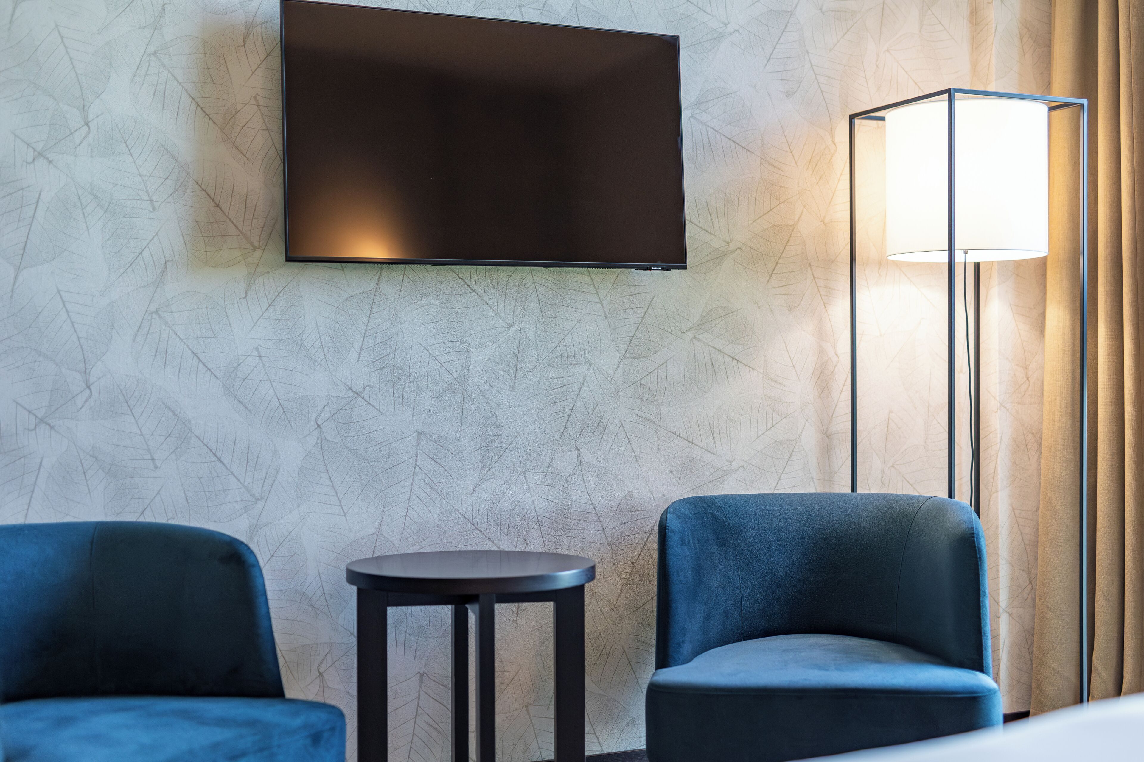 A hotel room with a flat-screen TV mounted on the wall, two blue armchairs, a small round table, and a floor lamp.