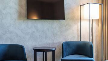 A hotel room with a flat-screen TV mounted on the wall, two blue armchairs, a small round table, and a floor lamp.