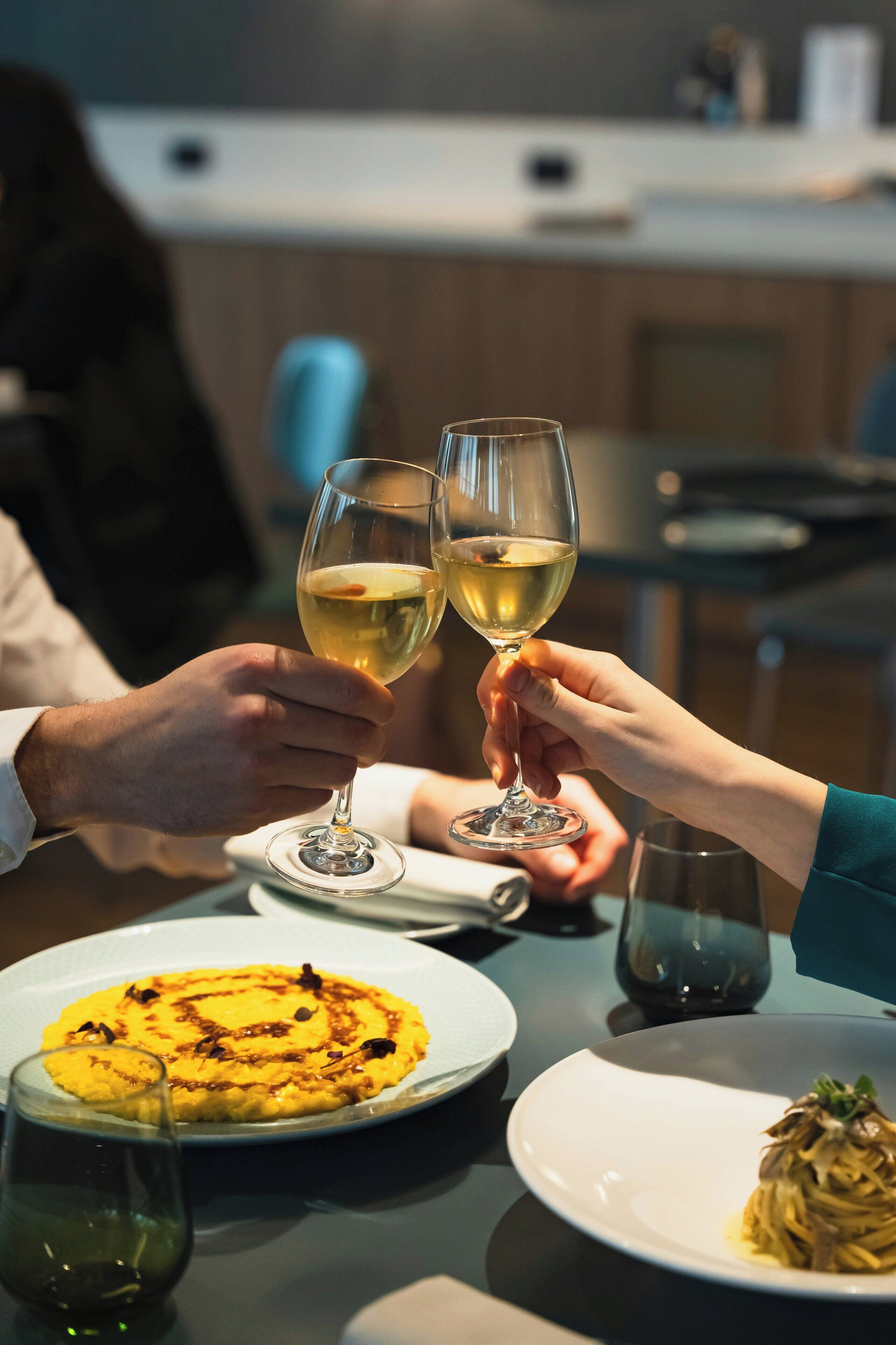 A dining table with plates of food, glasses of white wine, and a person toasting.