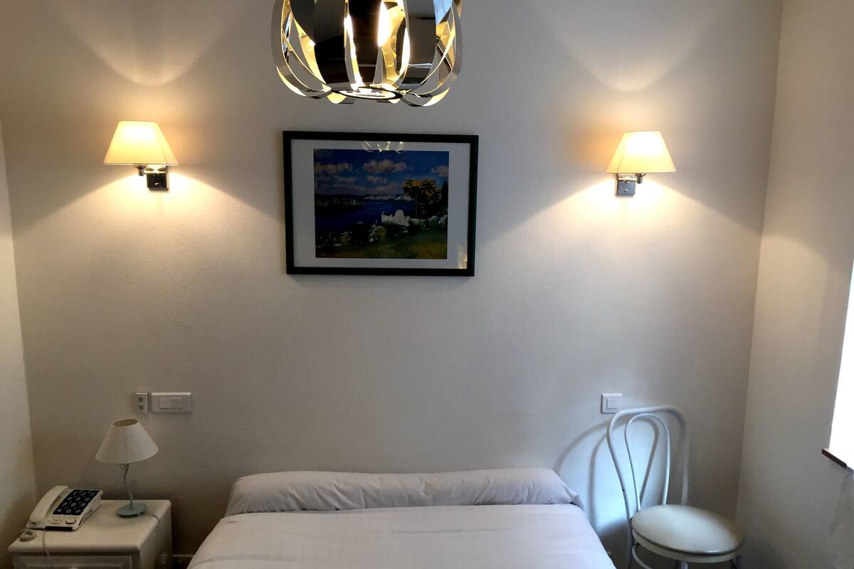 A hotel room with a bed, a nightstand with a lamp, a chair, and two wall-mounted lamps.