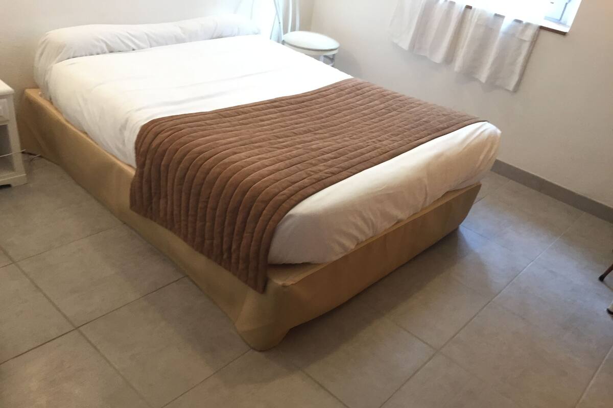 A neatly made bed with a brown blanket and white pillows in a room with tiled flooring and a small white stool.
