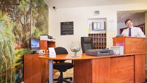 A reception desk with a computer, a glass of water, and a black office chair. There is a bookshelf with binders and a clock on the wall.