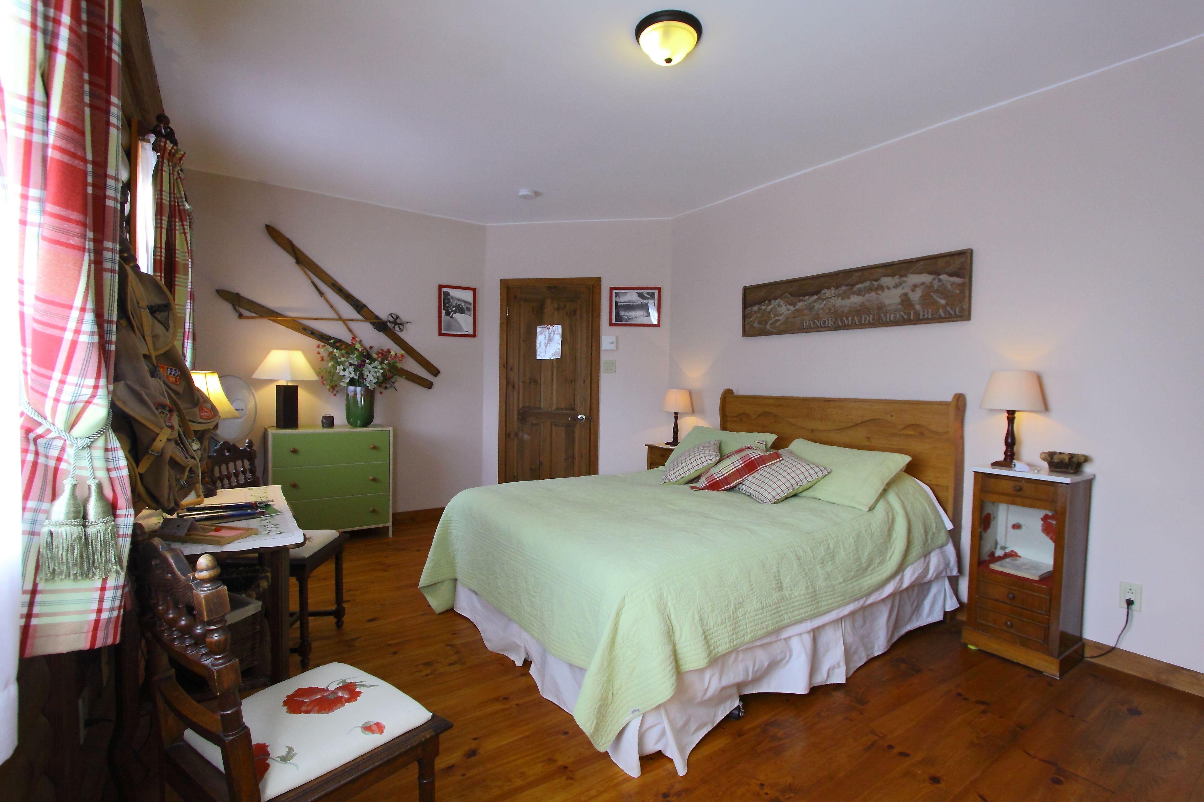 A bedroom with a wooden bed, green bedding, a green dresser, a wooden chair, a desk with a lamp, and a painting on the wall.