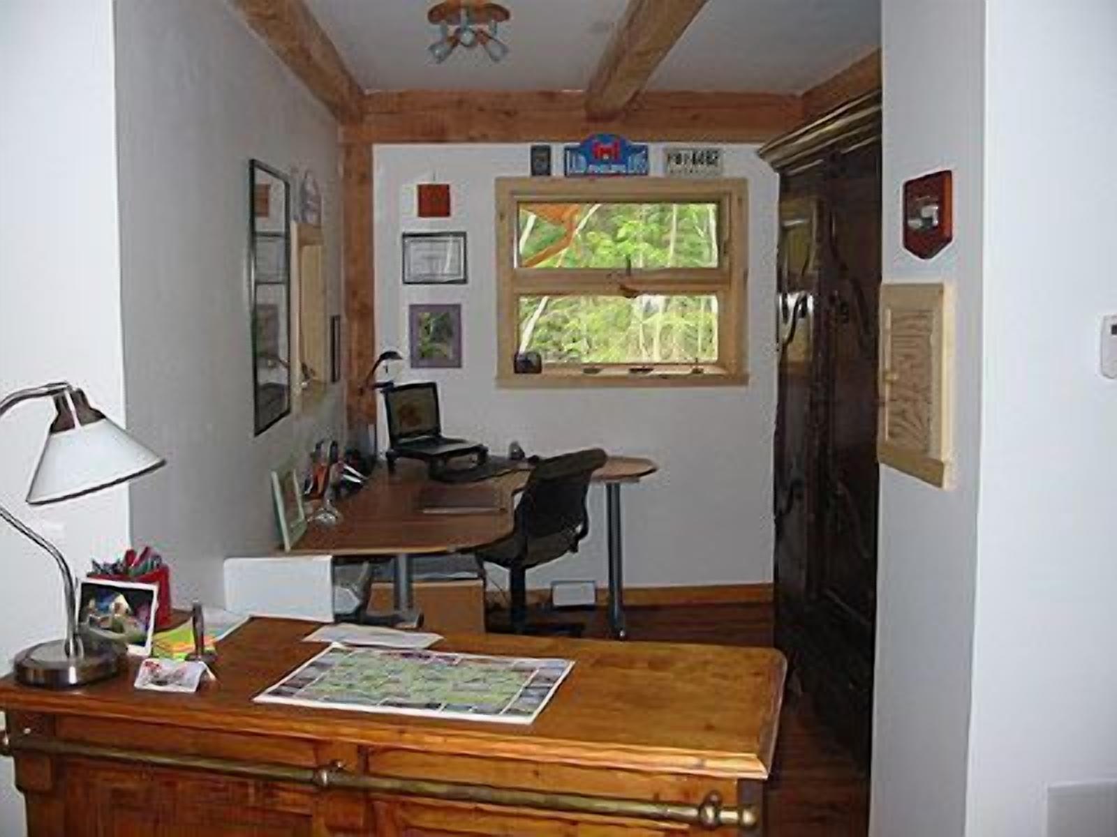A room with a wooden desk, a chair, a lamp, a map, and a window with a view of greenery.