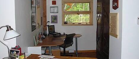 A room with a wooden desk, a chair, a lamp, a map, and a window with a view of greenery.