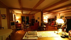 A cozy room with wooden beams, a dining table with newspapers, and various seating options including a red chair and a sofa. - Escale du Nord (Mont-Tremblant)
