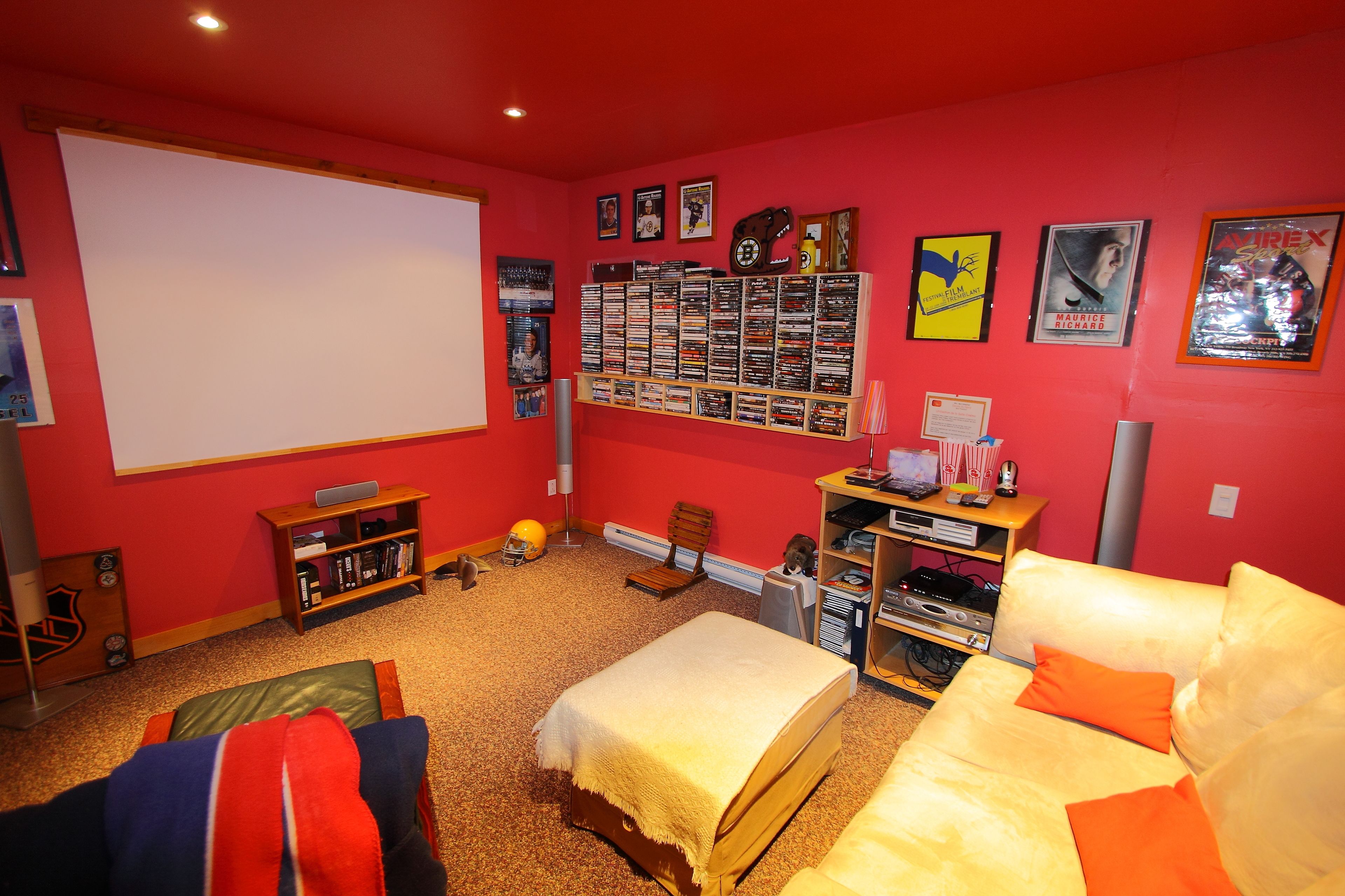 Una habitación con una cama grande, un sofá, un escritorio con una computadora, una estantería, una pantalla de proyector y varios carteles en la pared.