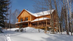 A wooden cabin with a large porch and snow-covered roof, surrounded by trees. - Escale du Nord (Mont-Tremblant)
