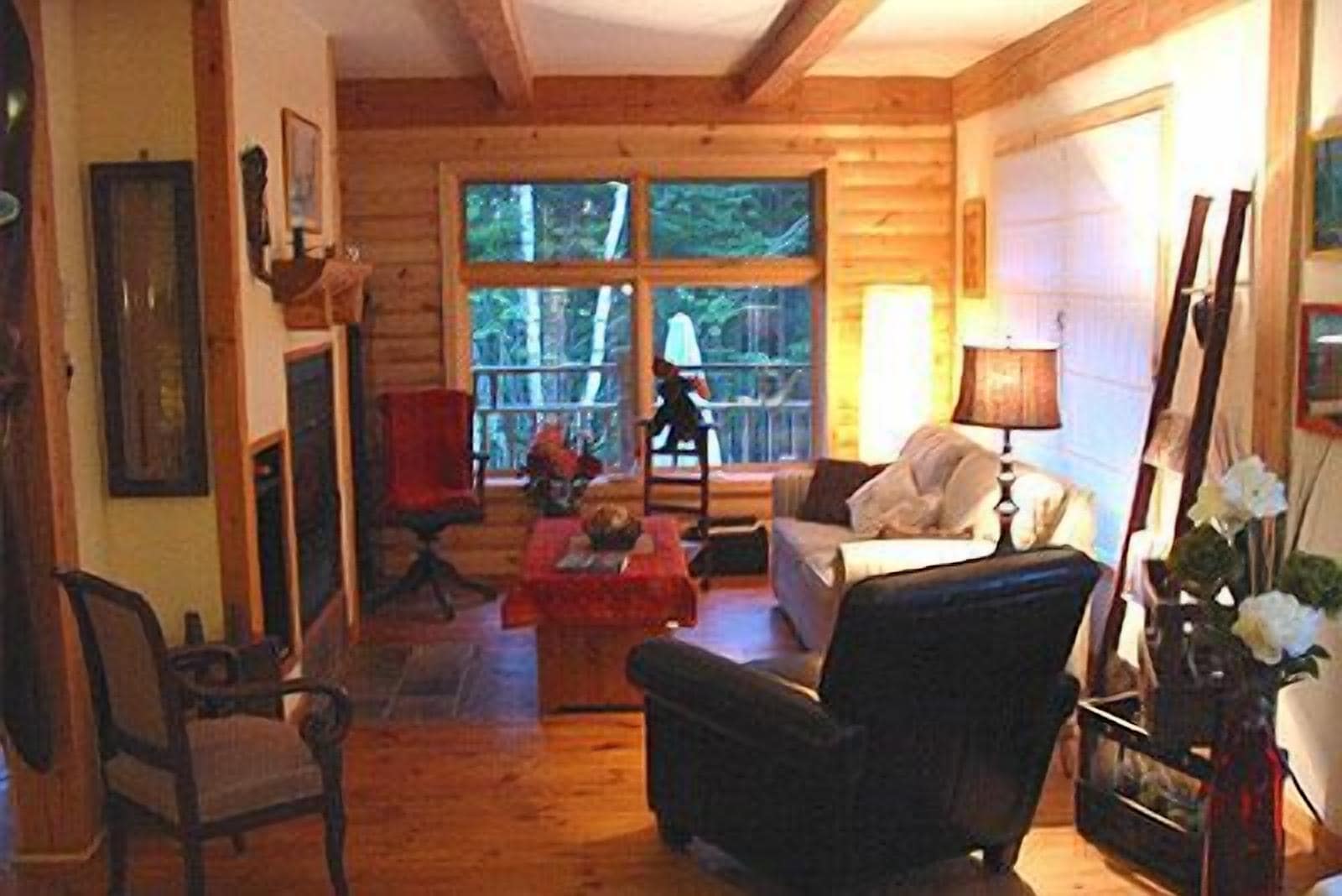 A cozy cabin interior with a wooden floor, a fireplace, a sofa, a chair, and a large window offering a view of the outdoors.