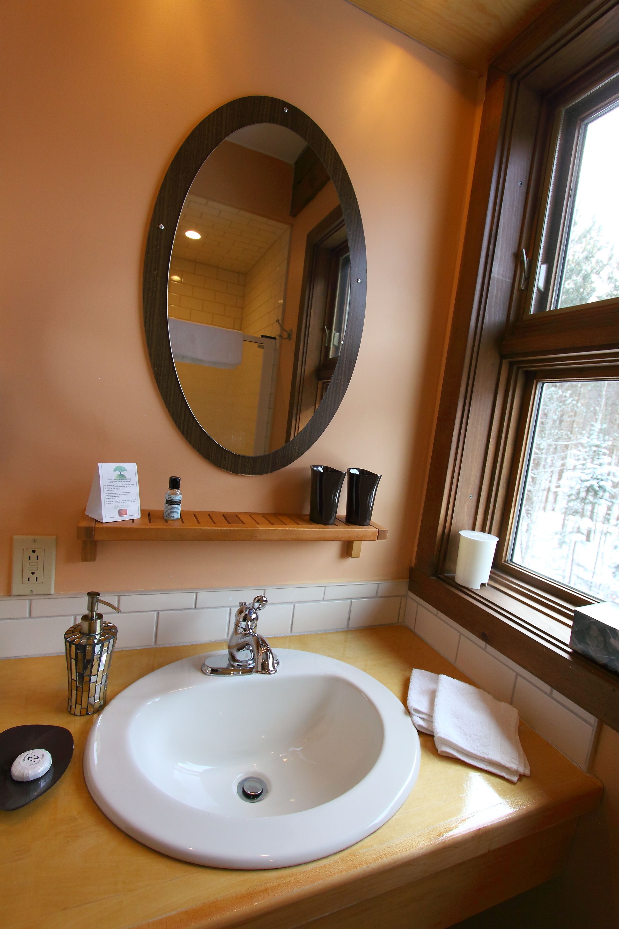 A bathroom with a white sink, a mirror, a window, and a wooden shelf with toiletries.