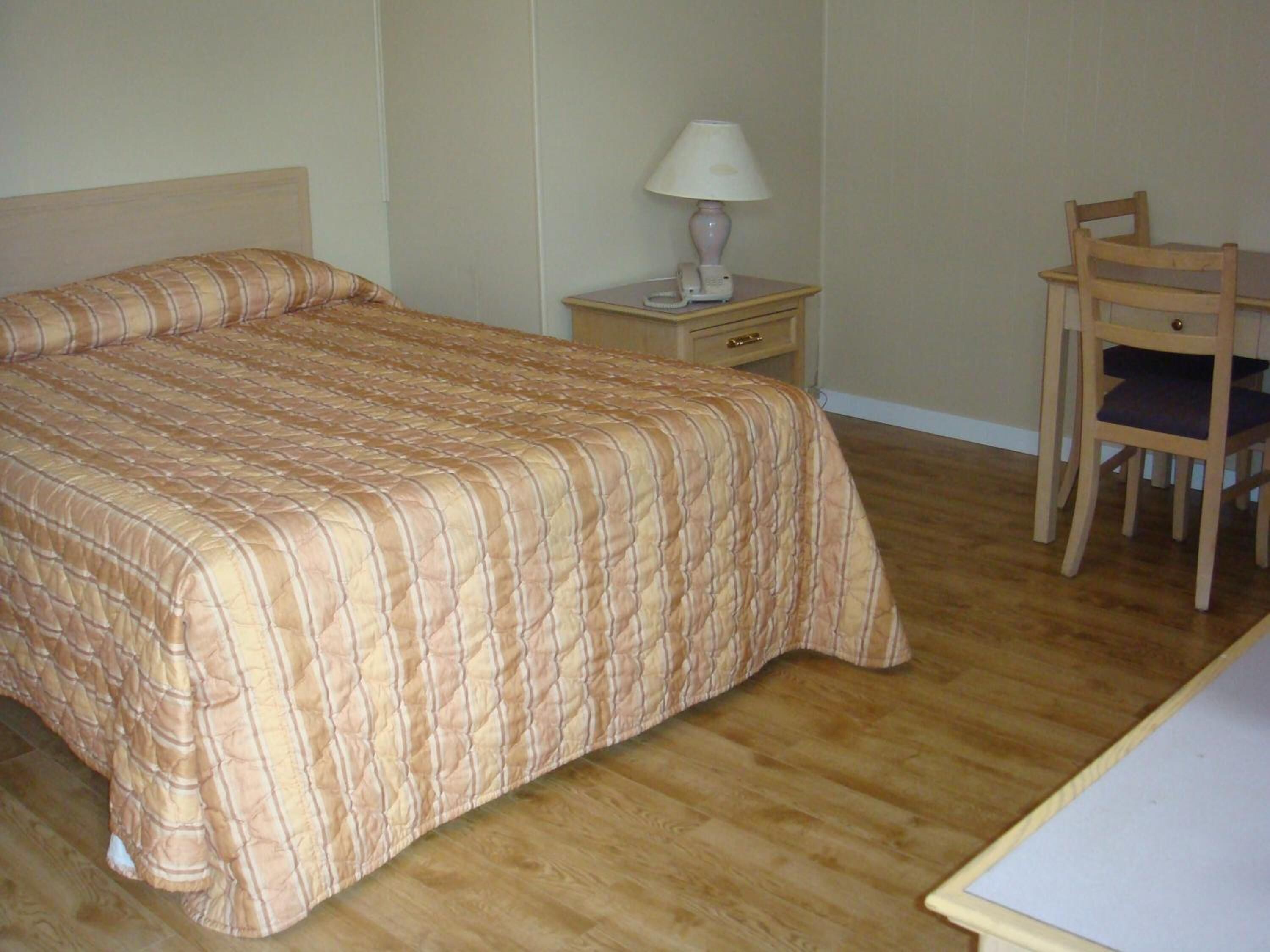 Un dormitorio con cama, una mesita de noche con lámpara, una silla y suelo de madera.