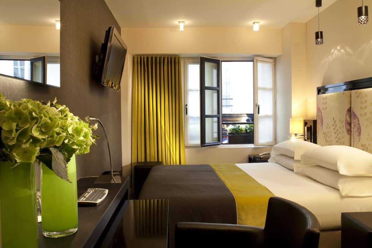 Ein Hotelzimmer mit einem Bett, einem Schreibtisch mit Computer, einem an der Wand befestigten Fernseher, einem Fenster mit Vorhängen und einer Vase mit Blumen.