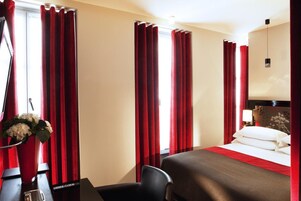 Hotellihuone, jossa on sänky, työpöytä tietokoneella, tuoli ja maljakko kukkineen.