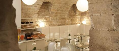 Un restaurante con paredes de piedra y mesas de vidrio, que ofrece opciones para el desayuno y sillas blancas.