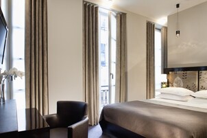 A hotel room with a bed, a desk, and a chair. There is a window with curtains, a door, and a view of a balcony.