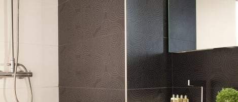 Un baño moderno con ducha, paredes de tono oscuro y estampado, un espejo grande y un lavamanos blanco con artículos de aseo.