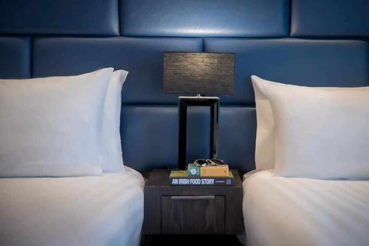 A hotel room with a blue headboard, two white pillows, a nightstand with a lamp, and a book titled 'An Irish Food Story.'