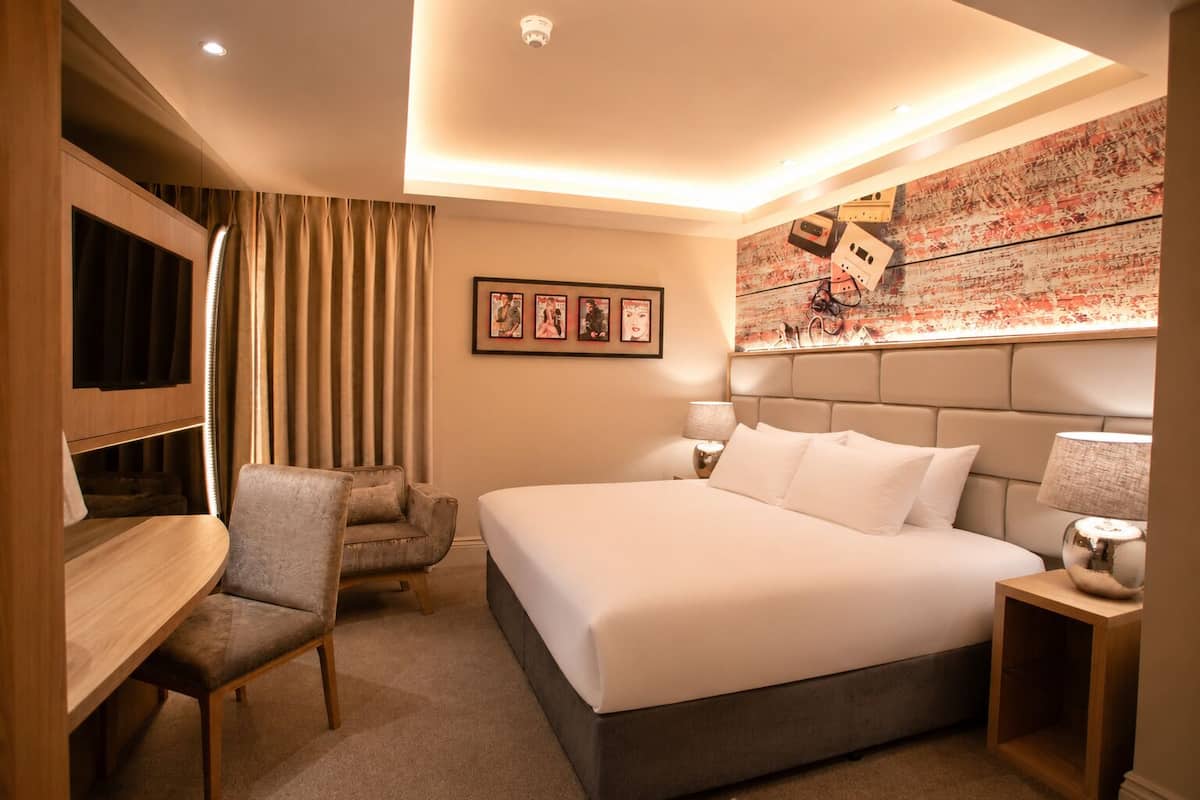 A hotel room with a bed, a desk, a chair, a headboard, and a wall with artwork.