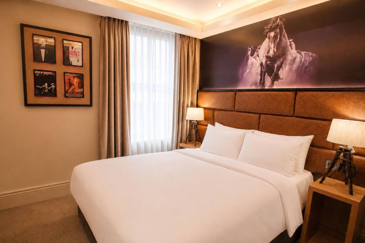 A hotel room with a large bed, two bedside tables with lamps, a large window with curtains, and a wall with framed movie posters.